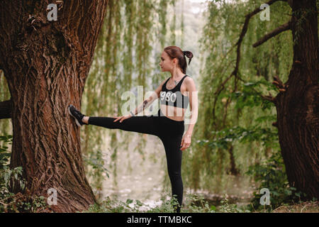 Female jogger exercising on tree in park by lake Stock Photo