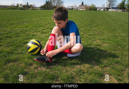 Full length of boy tying shoelace while sitting by soccer ball on grassy field Stock Photo