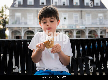 Cute boy eating ice cream cone while sitting on bench against building in city Stock Photo