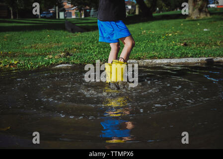 Low section of playful boy wearing rubber boots jumping in puddle at park Stock Photo