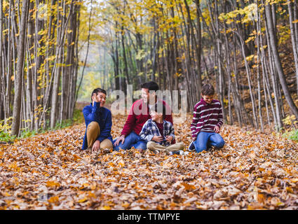 Happy father talking with sons while sitting on dry leaves in forest during autumn Stock Photo