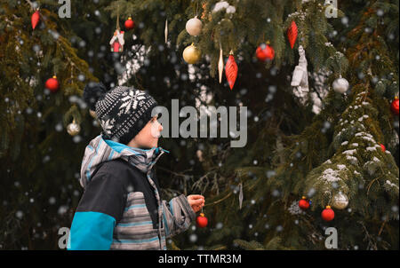 Young boy decorating tree outdoors with Christmas balls in the snow. Stock Photo