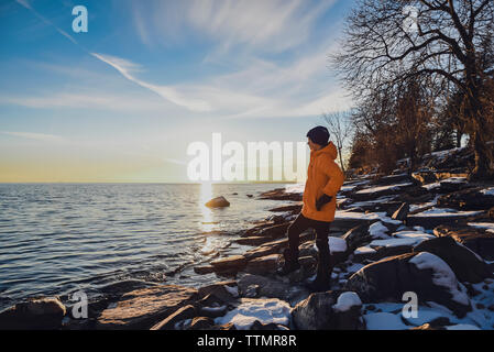 Boy standing alone on snowy rocks on lakeshore at sunset in winter. Stock Photo