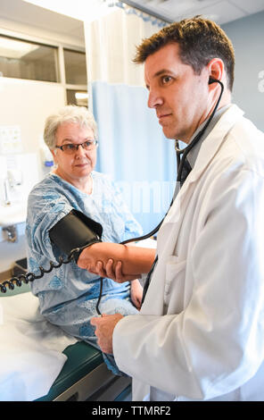 Doctor taking blood pressure of an older patient in gown in clinic. Stock Photo