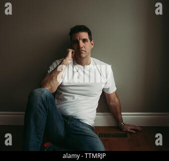 Man sitting on floor against plain wall resting face on his hand. Stock Photo
