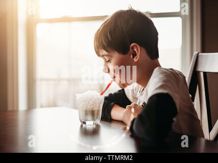 Happy boy blowing bubbles in glass of milk with a straw at the table. Stock Photo