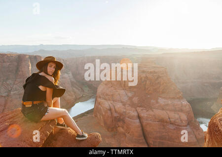 Full length portrait of young woman sitting on rock by Horseshoe Bend at desert during sunny day Stock Photo