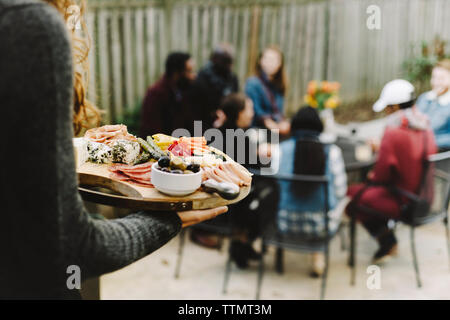 Woman carrying food on wooden board while friends enjoying in backyard Stock Photo