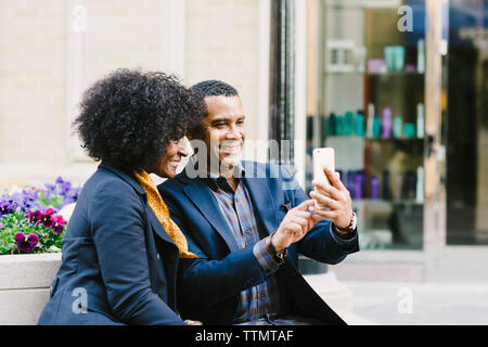Happy couple taking selfie through mobile phone while sitting on seat in city Stock Photo