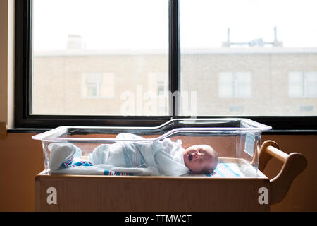 Baby boy yawning while sleeping in crib by window at hospital Stock Photo