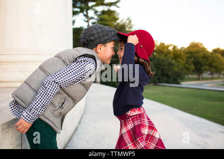 Side view of brother and sister looking at each other while standing on retaining wall in park during sunset Stock Photo