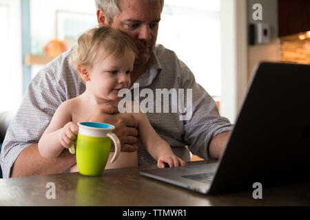 Dad Working From Home on Computer Holds Toddler With Green Sippy Cup Stock Photo