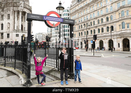 Three Siblings Pose in Front of Underground Sign on London Street Stock Photo