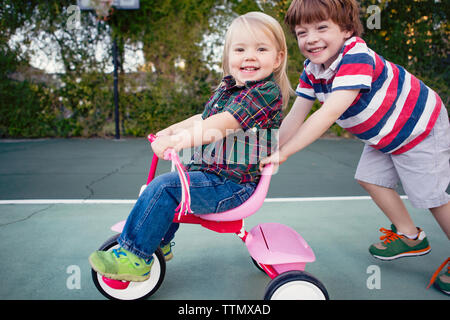 Portrait of happy boy pushing sister sitting on tricycle at yard Stock Photo