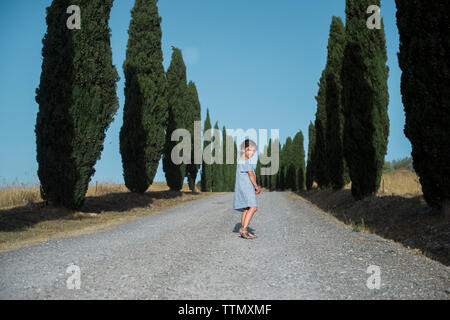 Portrait of girl standing on footpath amidst trees against clear blue sky during sunny day Stock Photo
