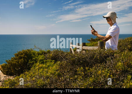 Man using phone while standing on cliff by sea against sky Stock Photo
