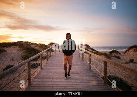 Smiling hiker walking on footbridge at beach against sky during sunset Stock Photo
