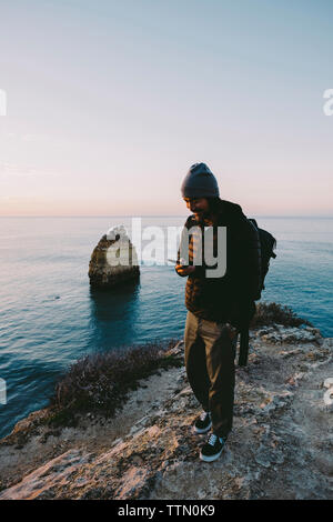 Man using smart phone while standing on rock formation against sky during sunset Stock Photo