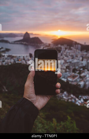 Cropped hand of man photographing city by sea against cloudy sky during sunset Stock Photo
