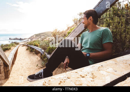 Smiling hiker looking away while sitting against railing at beach Stock Photo