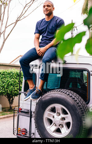 Full length of smiling young man sitting on SUV's rooftop Stock Photo