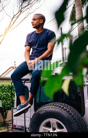 Full length of young man looking away while sitting on SUV's rooftop Stock Photo
