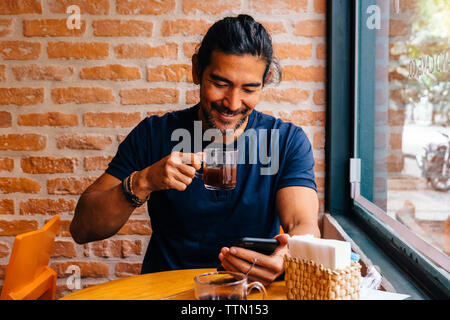 Smiling mature man with black coffee using smart phone against brick wall in cafe Stock Photo