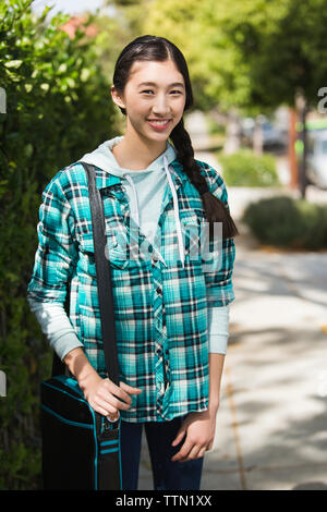 Portrait of cheerful teenage girl carrying shoulder bag while standing against plants at park Stock Photo