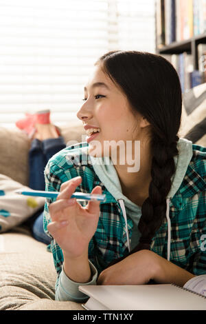 Smiling teenage girl looking away while doing homework on couch at home Stock Photo