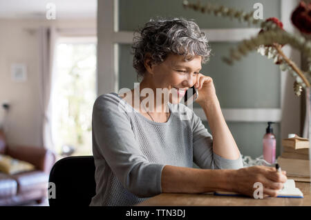Mature woman talking on smart phone and writing in book at home Stock Photo