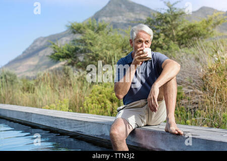Mature man drinking coffee while sitting by swimming pool against mountain Stock Photo