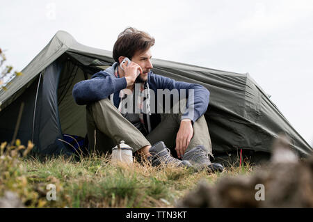 Man answering smart phone outside tent on hill against clear sky Stock Photo