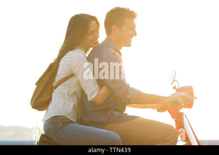 Side view of couple riding on motor scooter against clear sky on sunny day Stock Photo
