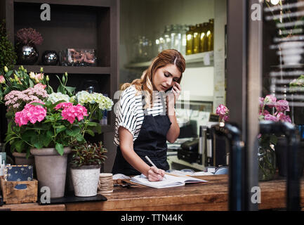 Florist writing in note pad while talking on mobile phone Stock Photo