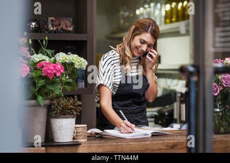 Florist writing in note pad while using mobile phone Stock Photo