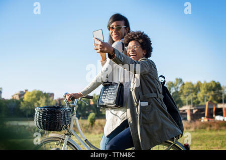 Happy friends taking selfie through phone while riding bicycle on field Stock Photo
