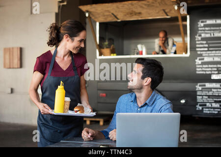 Female owner serving food for customer sitting at table on street against food truck Stock Photo