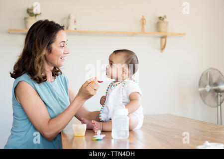 Mother feeding baby boy sitting on table at home Stock Photo