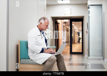 Senior doctor using laptop computer while sitting on sofa in corridor Stock Photo