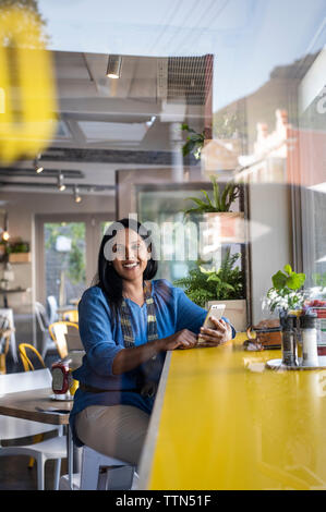 Smiling woman using smart phone while sitting at table in cafeteria Stock Photo