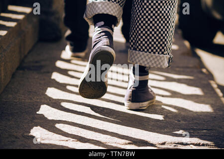 A man in sneakers and fashionable plaid pants walking on the stone pavement, which falls striped shadow of the fence Stock Photo