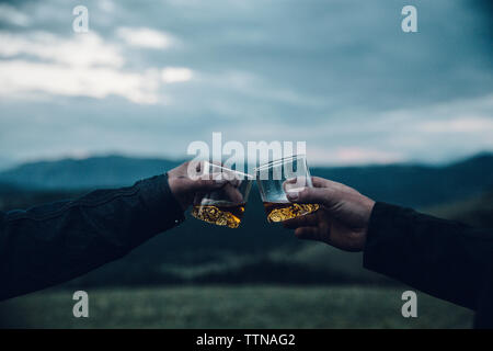 Cropped hands of friends toasting drinks on mountain against cloudy sky during sunset Stock Photo