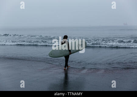 Rear view of woman with surfboard walking at beach against sky Stock Photo