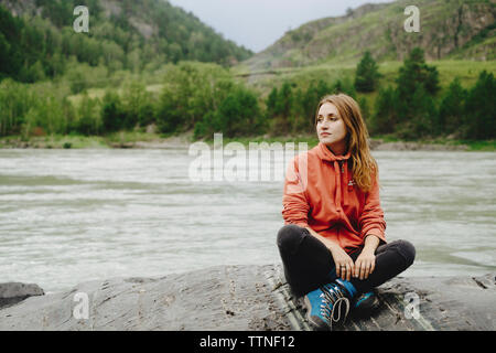 Thoughtful female hiker relaxing on rock at riverbank against mountain Stock Photo