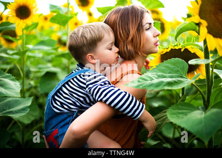 Woman standing in the sunflower field, holding her son on her back.