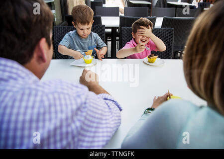 Happy boy covering eyes while eating desert with family in restaurant Stock Photo