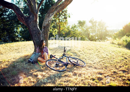 Young woman leaning on tree trunk while sitting on grassy field Stock Photo