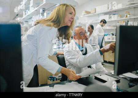 Doctors discussing at desk over desktop computer while coworkers working in background at hospital Stock Photo