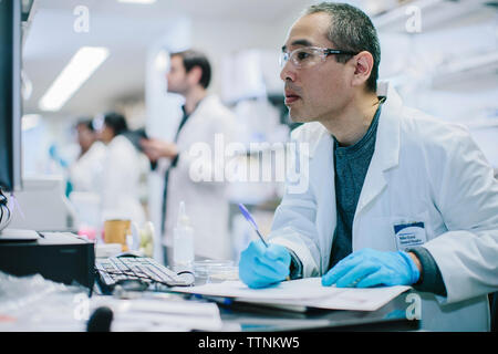 Male doctor writing at desk while coworkers working in background Stock Photo
