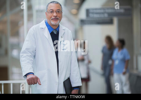 Portrait of male doctor standing in corridor while female coworkers discussing in background Stock Photo